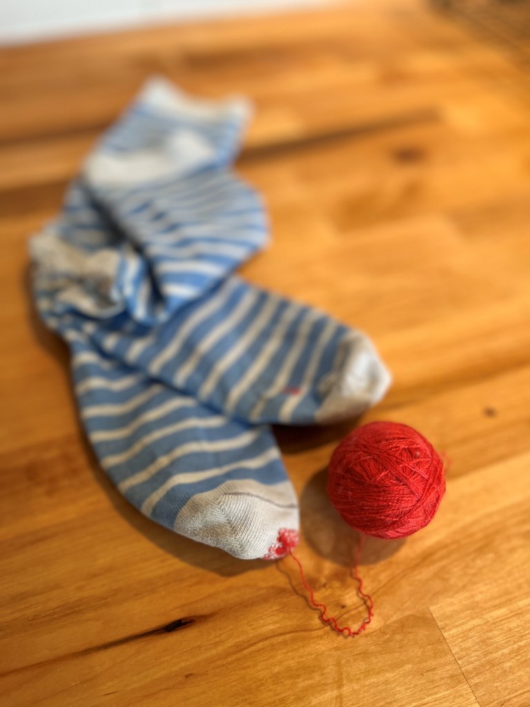 Une balle de fil rouge, ainsi qu'une paire de bas dont un trou à l'orteil a été réparé avec le fil provenant de la balle montrée sur l'image